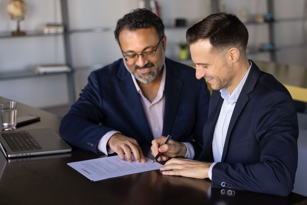 Two multiethnic businessmen finalizing formal meeting, signing contract with smile, looking satisfied and contented with partnership
