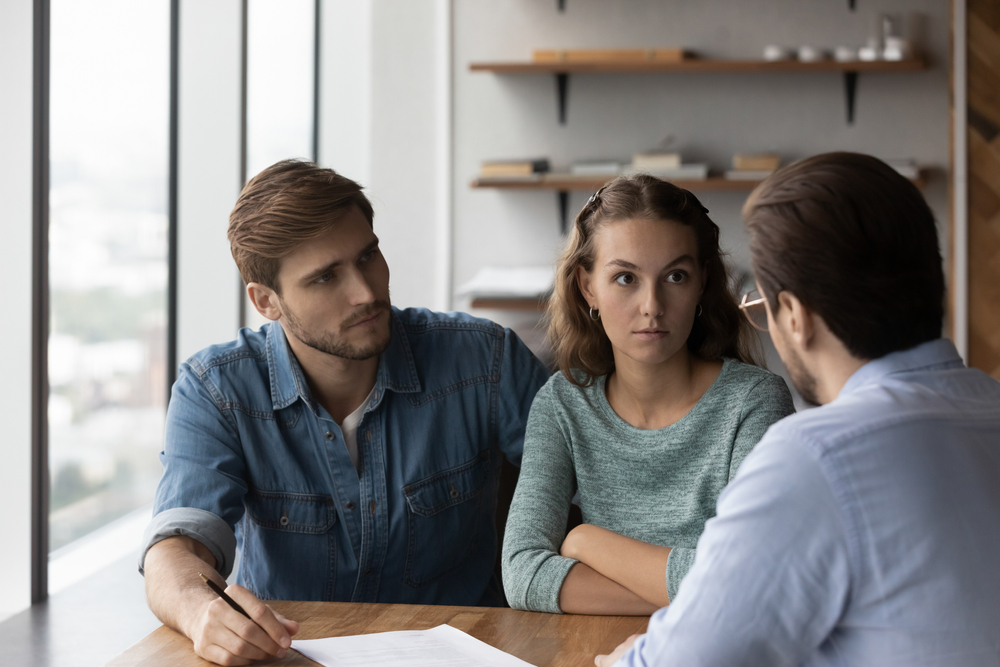 Unhappy young Caucasian couple buyers talk with male realtor or real estate agent dissatisfied with contract terms regulations